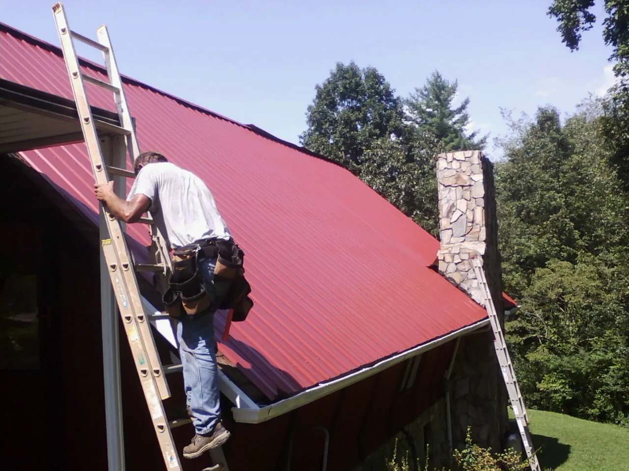 Roofer on a ladder beside a red metal roof and stone chimney — Soffit & Fascia Repair by Roofing Brothers in Kershaw